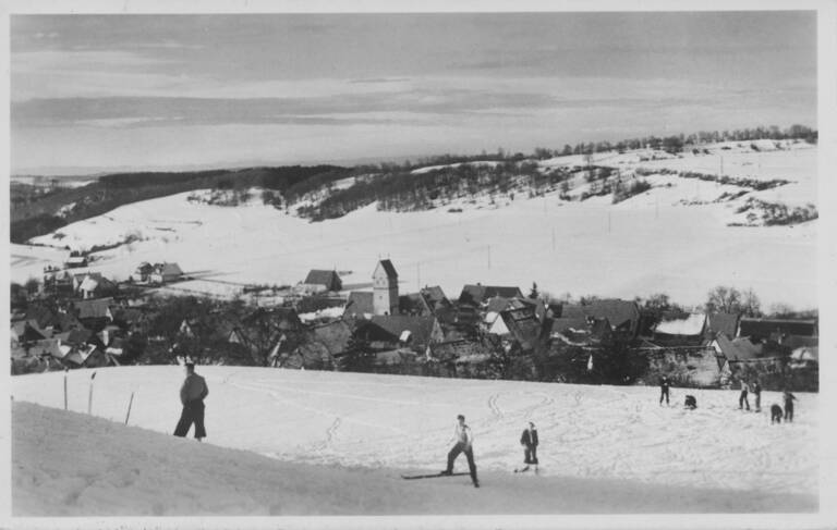 Historisches Schwarz-weiß-Foto von Skifahrern. Im Hintergrund ist die Ortschaft Trailfingen erkennbar